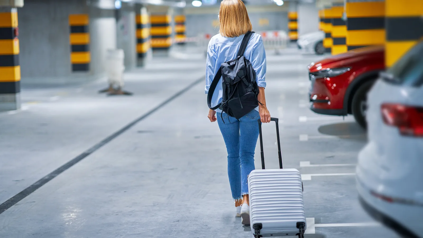 Kobieta z walizką i plecakiem idzie między autami po podziemnym parkingu