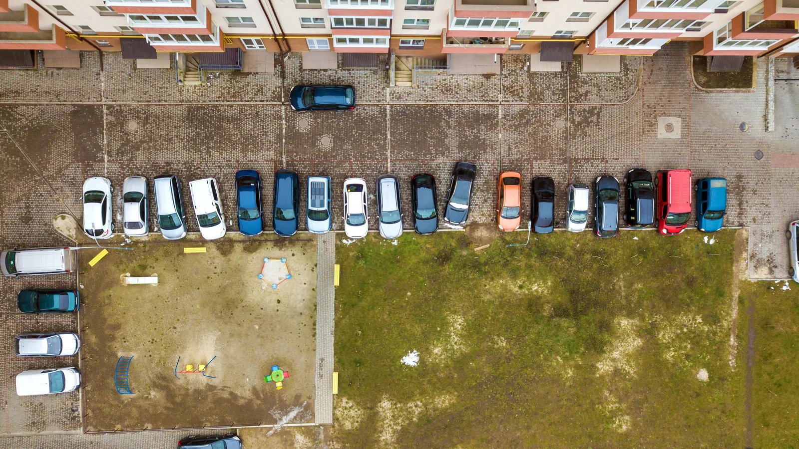 Kadr z drona na rząd zaparkowanych aut przy bloku i plac zabaw obok